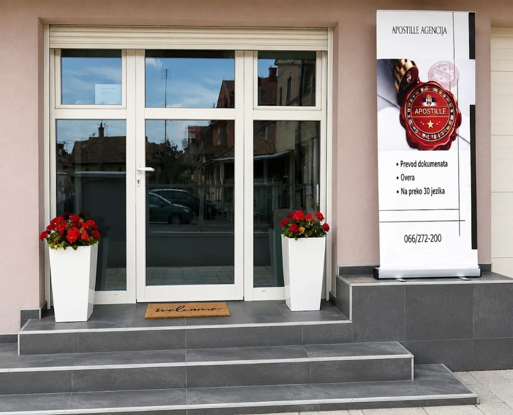 Ulaz kancelarije u Šapcu: staklena vrata, saksije sa crvenim cvećem i roll-up baner agencije.